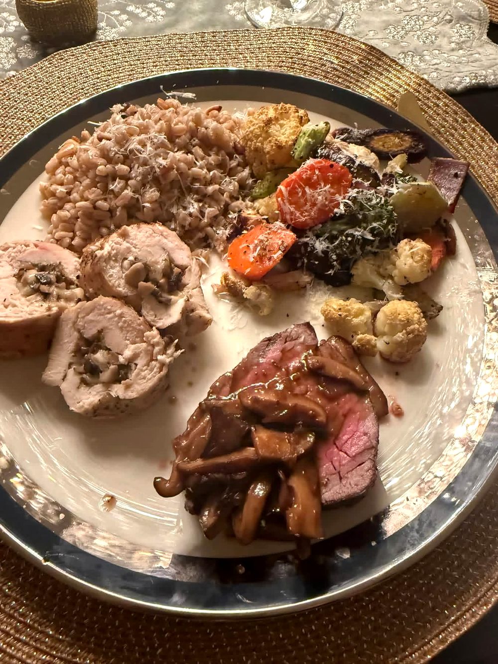 A plated dinner at a candlelit table: sliced beef tenderloin with mushrooms, a stuffed chicken roulade, farro with pine nuts, and roasted vegetables