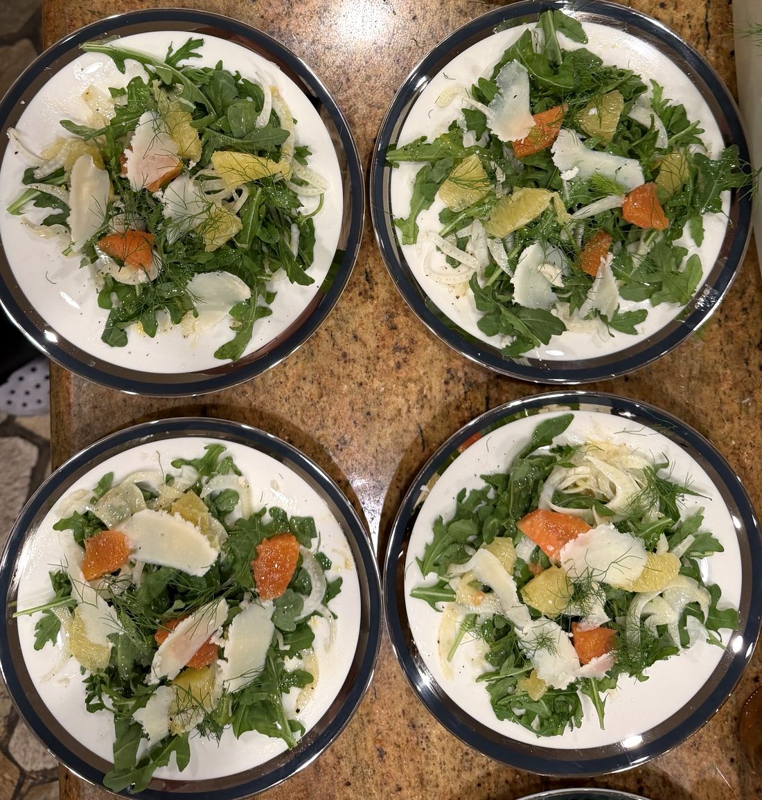Four plates of fennel, orange and arugula salad with shaved Parmesan, lined up on a granite counter ready to serve